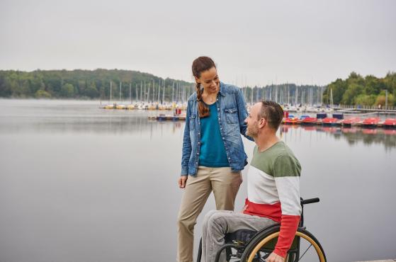Mann im Rollstuhl und Frau spazieren am Ufer eines Sees